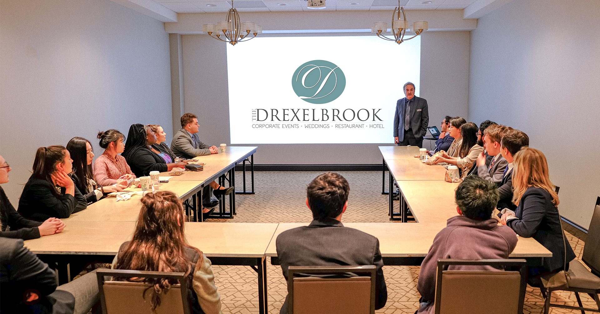 About a dozen young people sitting around a u shaped table looking at a screen projected onto the wall of a small break out room.