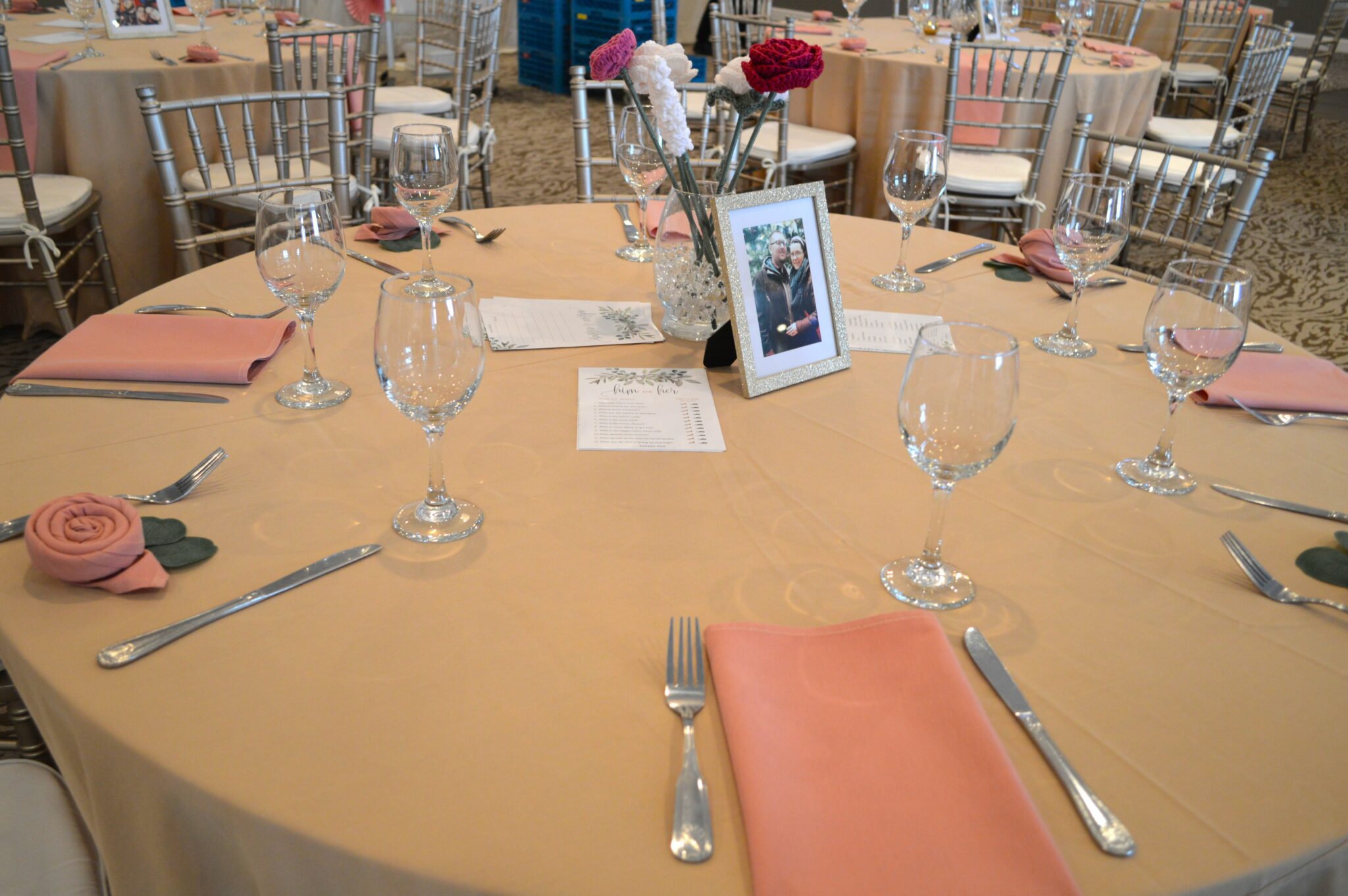 Round table set up with an ivory tablecloth and pink linens. napkins are folded in the shape of roses and centerpieces include crocheted flowers. 