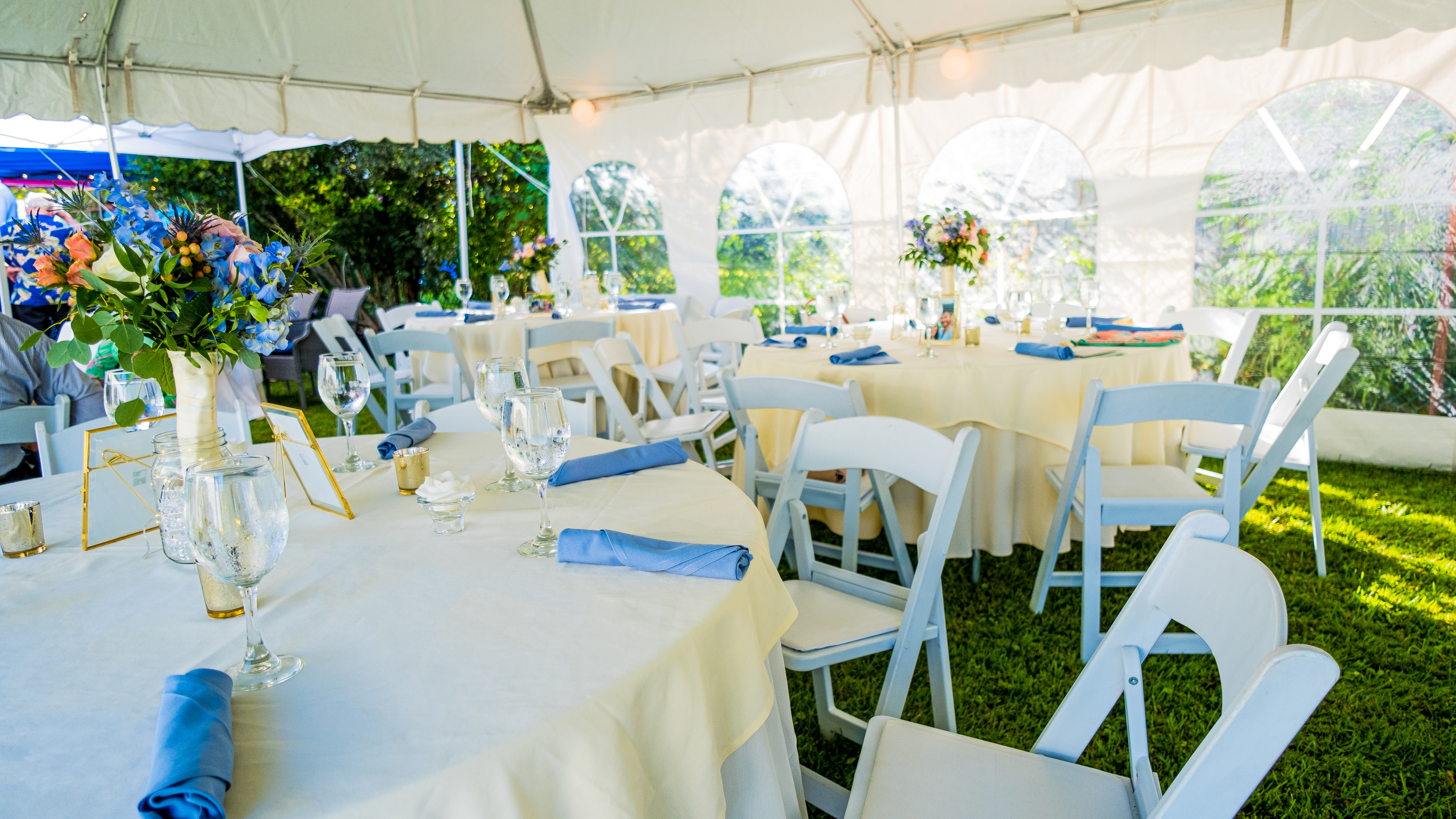 An outdoor wedding reception set up underneath a white tent. Tables with ivory linens are set up with white chairs. Blue napkins and blue flower arrangements are on the tables. 