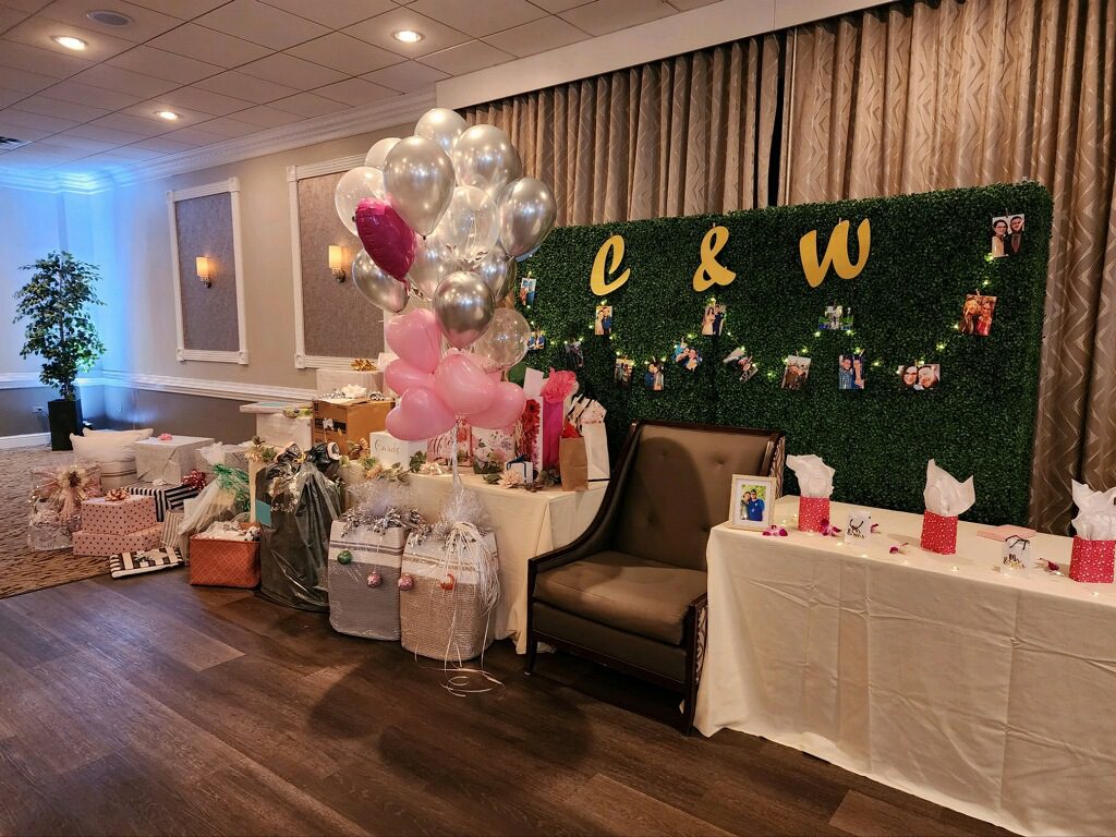 Bridal shower set up in a ballroom at The Drexelbrook. Presents and balloons are surrounding a chair set up infront of a wall of greenery displaying photos of the marrying couple.